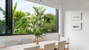 A dining area with a wooden table, white chairs, a vase with green plants, and a window with a view of tropical plants. - Pacific Oasis l Private Pool, 450mtrs to Beach (Coolum Beach)