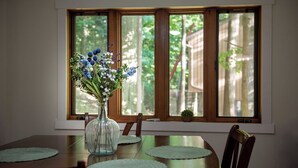 A dining table with a vase of flowers, a window with a view of trees, and a small potted plant on the windowsill. - Em's Lake House - Newly Renovated 5 Bedroom Lake House (Sterling)