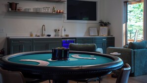A room with a green felt table, chairs, a television mounted on the wall, and a kitchen area with cabinets and a sink. - Em's Lake House - Newly Renovated 5 Bedroom Lake House (Sterling)