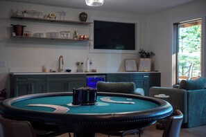 Ein Zimmer mit einem grünen Filztisch, Stühlen, einem an der Wand befestigten Fernseher und einer Küchenzeile mit Schränken und einer Spüle.