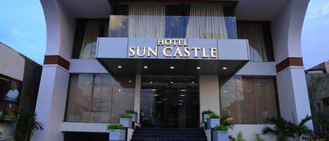 L’entrée d’un hôtel, avec une enseigne indiquant « HÔTEL SUN CASTLE », un escalier menant à l’étage et des plantes en pot à sa base.