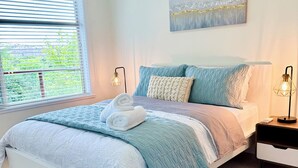 Un dormitorio con cama, mesitas de noche, una ventana con persianas y una obra de arte colgada en la pared.