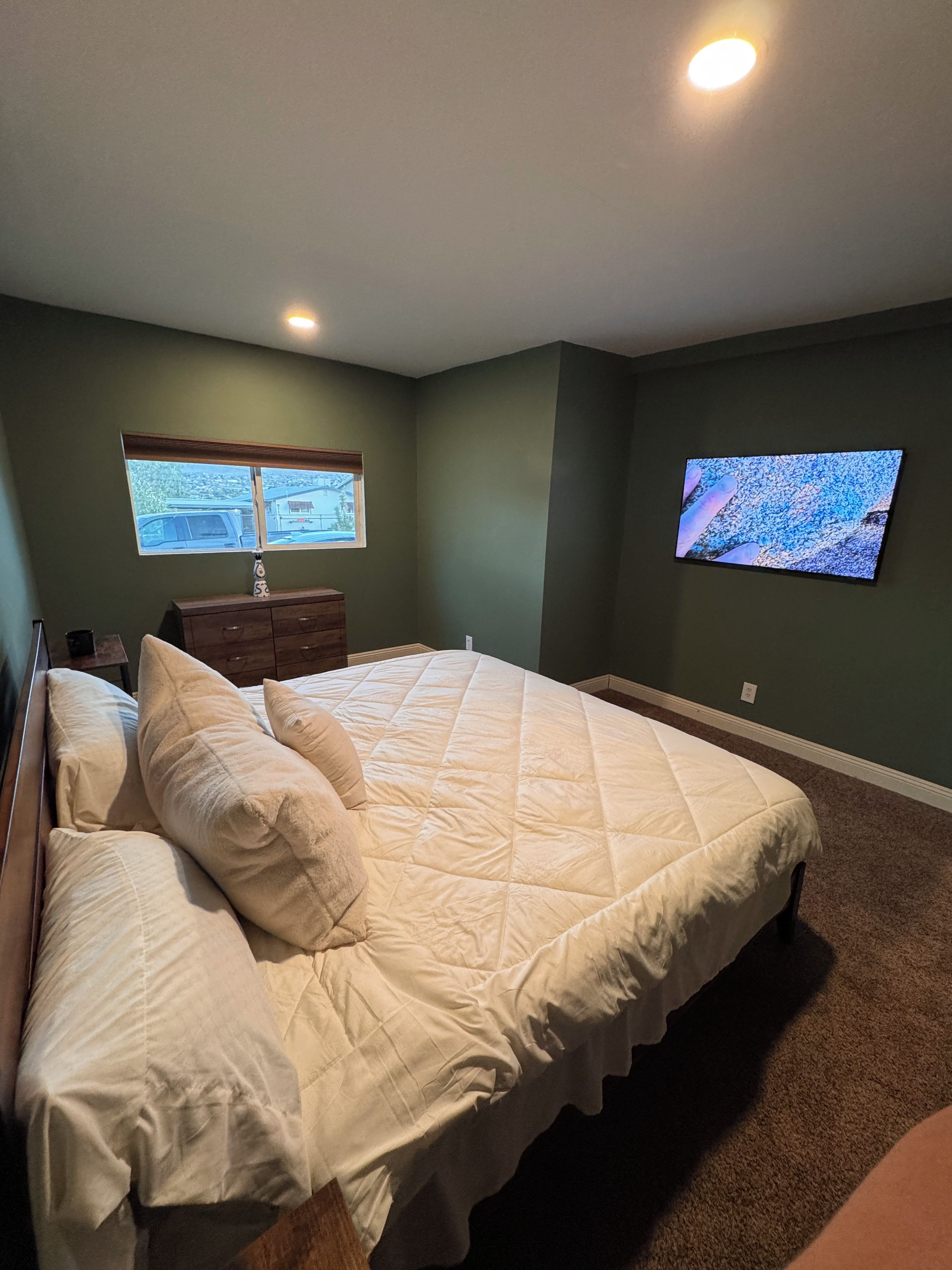 A bedroom with a large bed, a dresser, a television on the wall, and a window with a view of the outside.