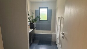Un couloir étroit mène à une salle de bain dotée d’une fenêtre, d’une plante en pot et d’un tapis à carreaux.