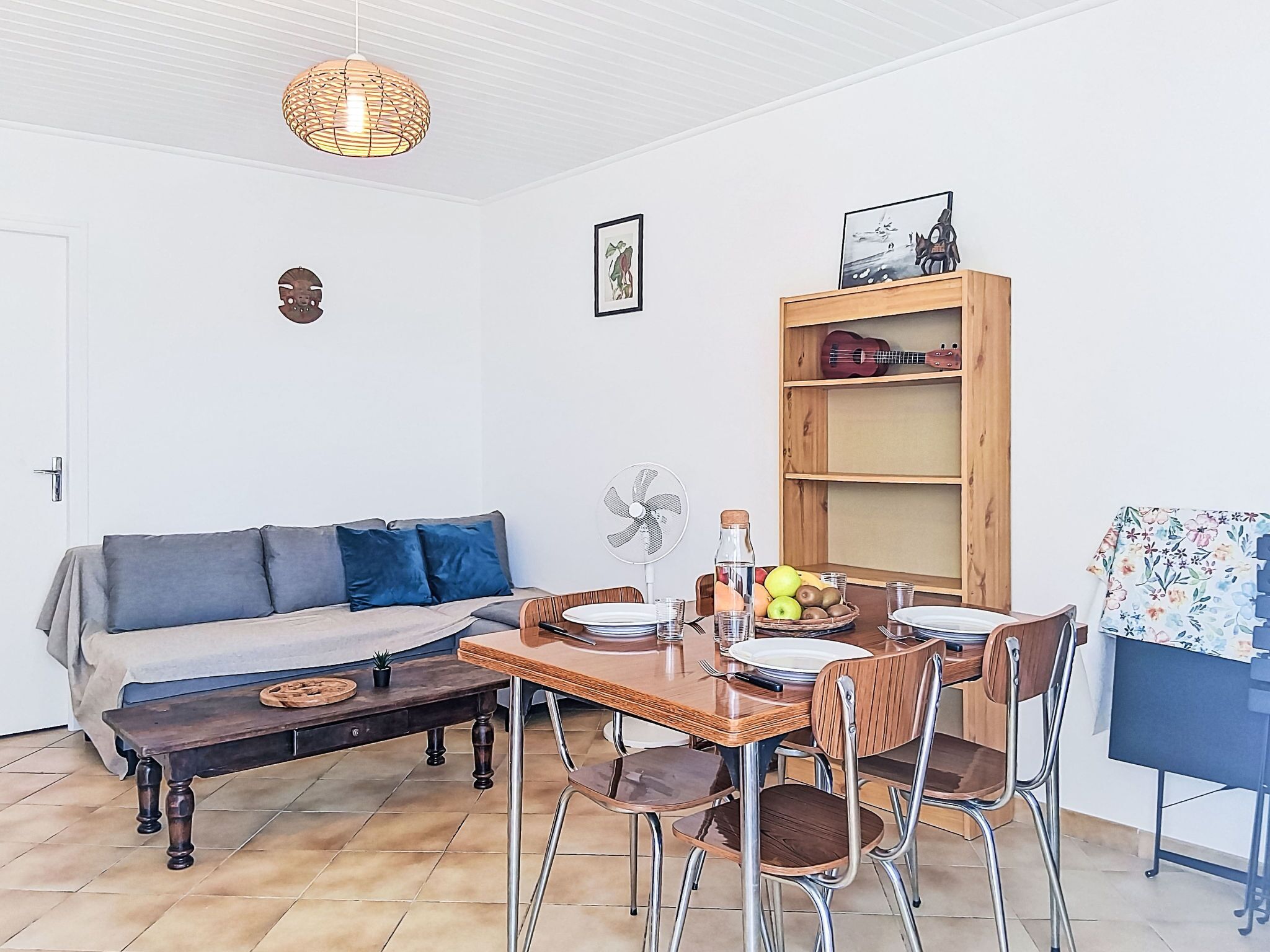 A dining area with a wooden table, chairs, a sofa, a fan, and a shelf with a guitar.