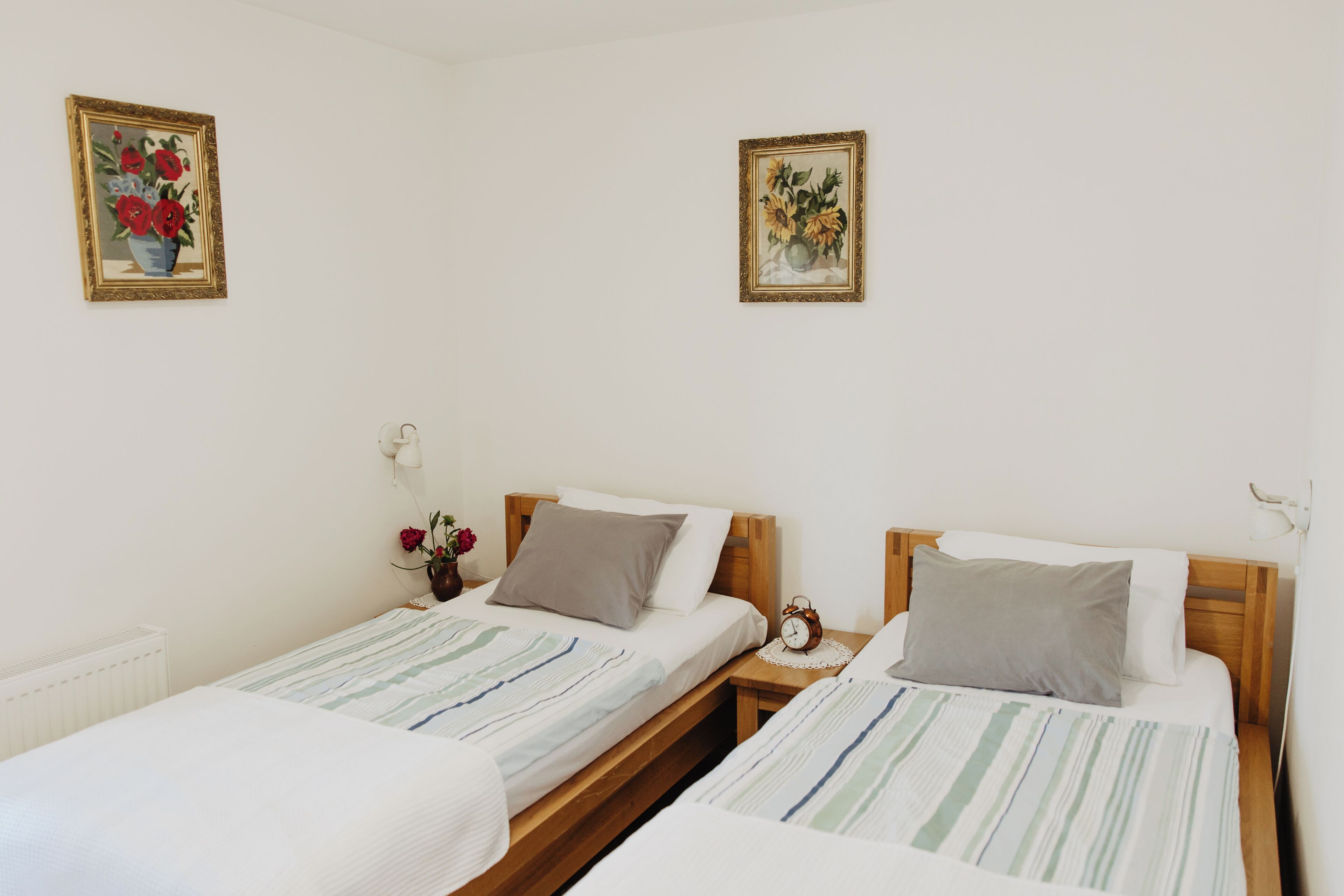 Une chambre avec deux lits simples, une table de chevet avec une horloge, un vase avec des fleurs et deux tableaux encadrés au mur.