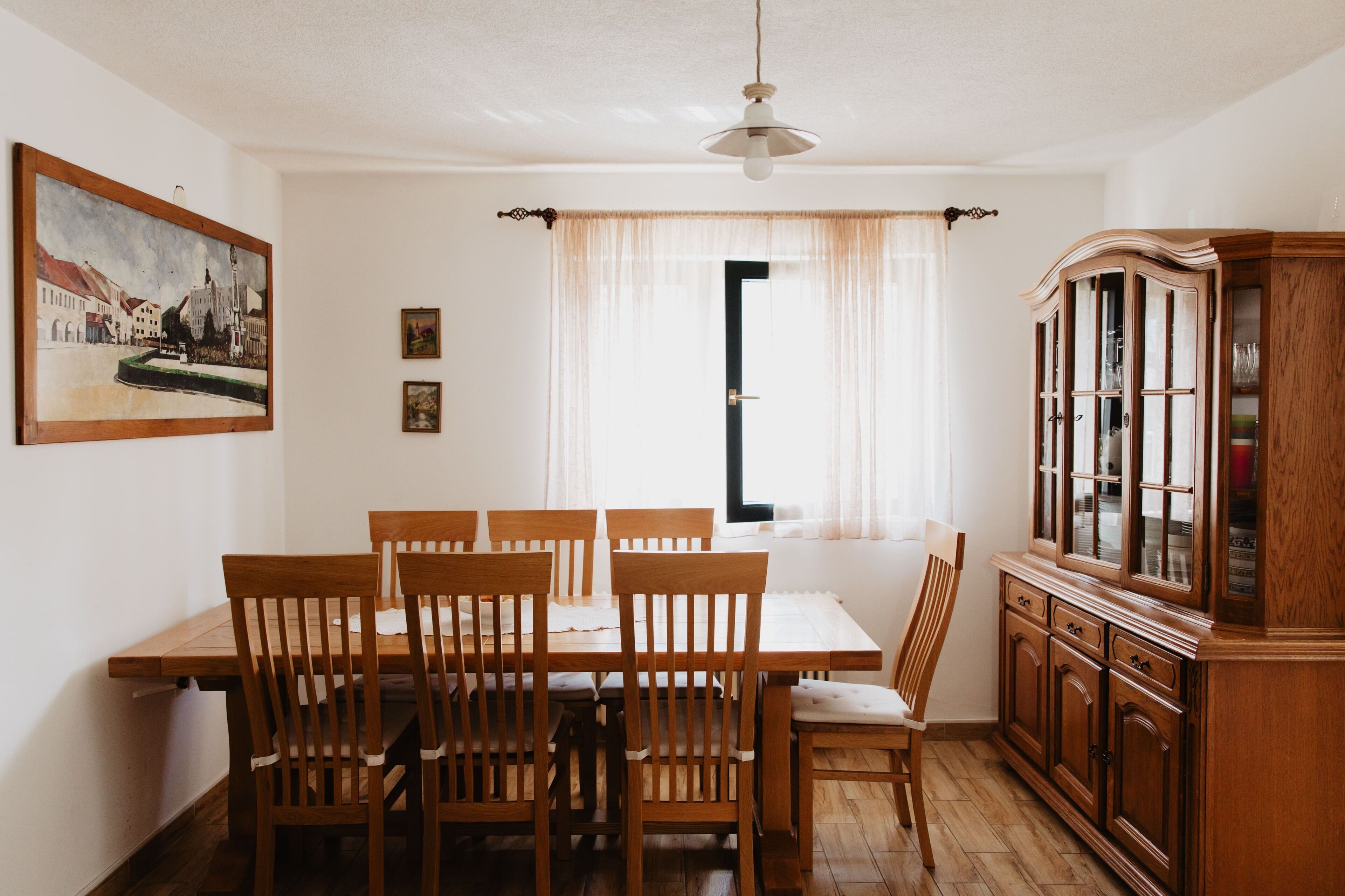 Un comedor con una mesa y sillas de madera, un aparador con puertas de vidrio y un cuadro en la pared.