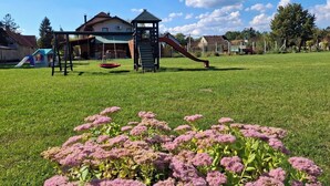 A playground with swings and slides, surrounded by a grassy field and houses in the background.