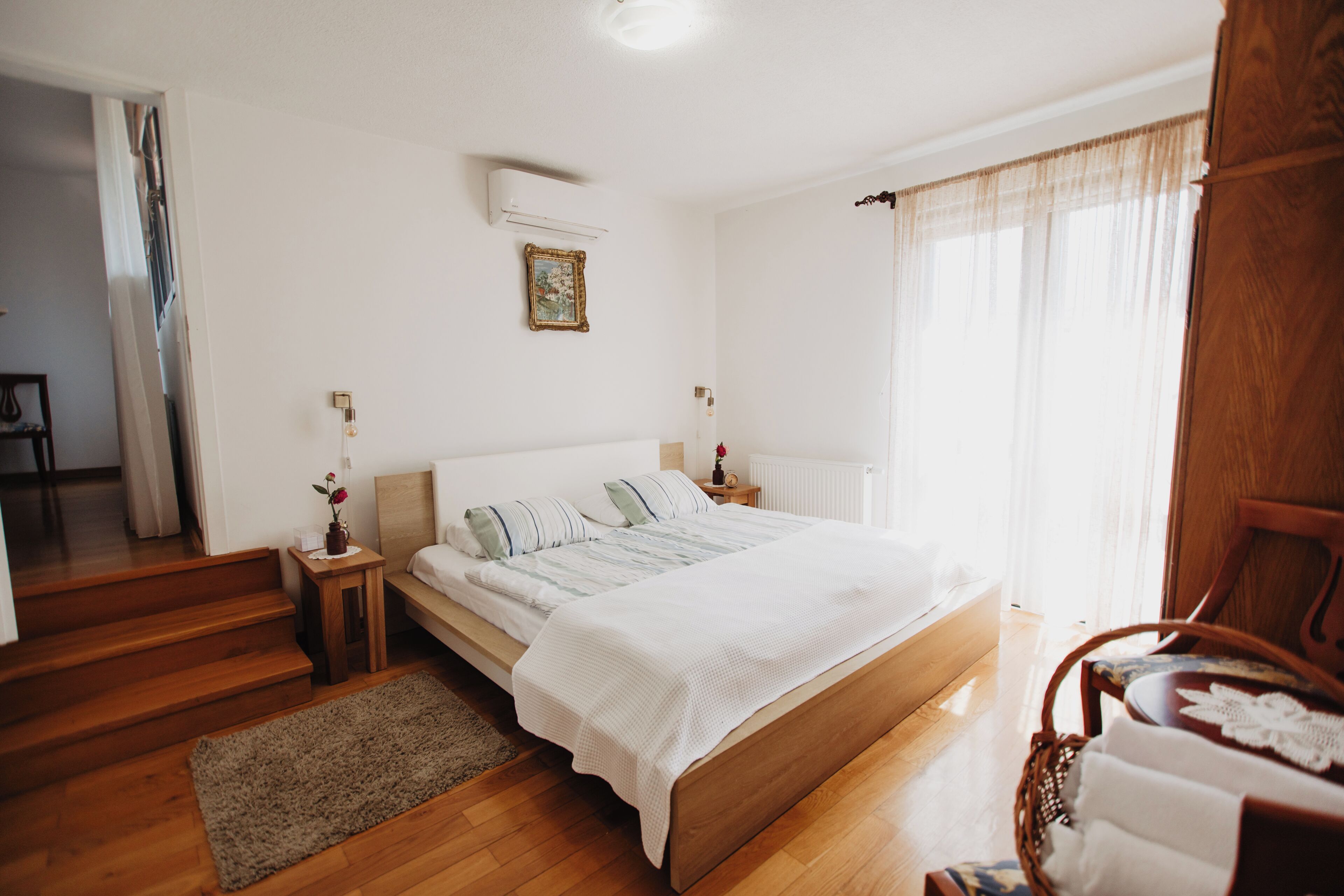 Une chambre avec un lit en bois, une table de chevet ornée d’un vase, une commode, une fenêtre avec des rideaux légers et un plancher en bois.