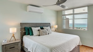 A bedroom with a bed, nightstands, a ceiling fan, an air conditioner, a window with a view, and a lamp.