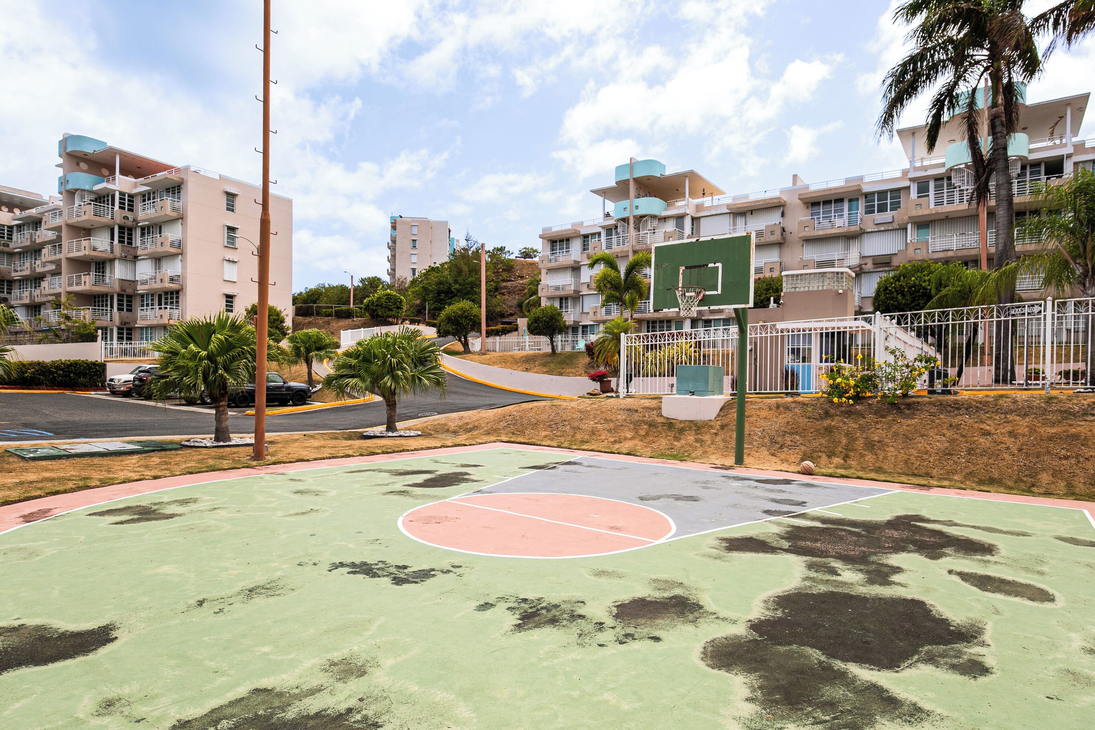 Un terrain de basket avec une surface verte et rouge, entouré d’une clôture, avec des immeubles d’habitation en arrière-plan.