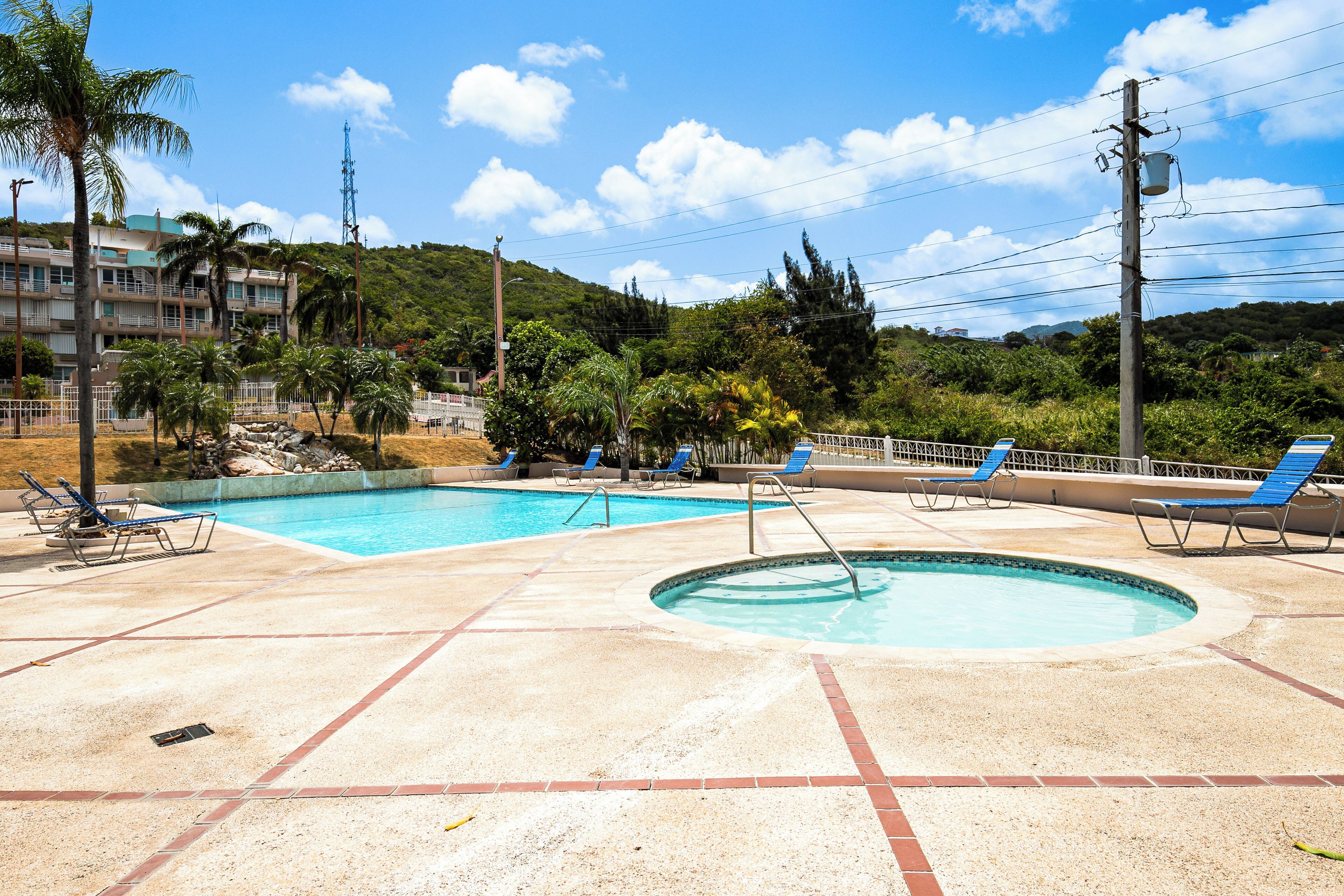 Une piscine entourée de chaises longues bleues, un petit jacuzzi rond et une vue sur une colline avec des bâtiments en arrière-plan.