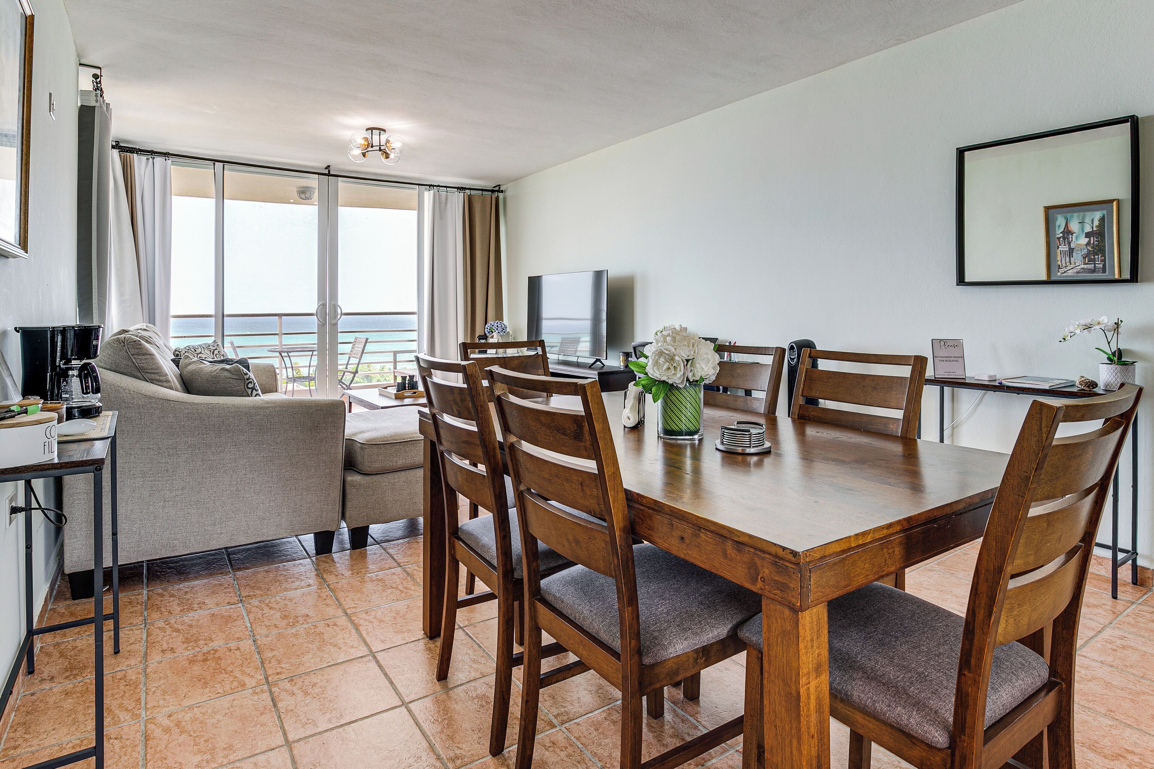 Un comedor con mesa y sillas de madera, televisor y vista al mar a través de amplios ventanales.