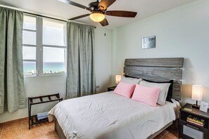 Ein Schlafzimmer mit einem Bett, einem Nachttisch, einem Deckenventilator, einem Fenster mit Vorhängen und Blick aufs Meer.