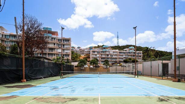 An outdoor tennis court with a net, surrounded by a fence, and buildings in the background.