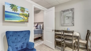 Una camera con un letto, una poltrona blu, un tavolo da pranzo con sedie, un quadro raffigurante una spiaggia e uno specchio appeso al muro.