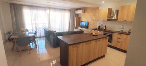 A modern kitchen with wooden cabinets, a central island, and a dining area with a sofa and chairs. - GrandBlue Vista Premier Seafront Apartment (Żebbuġ)