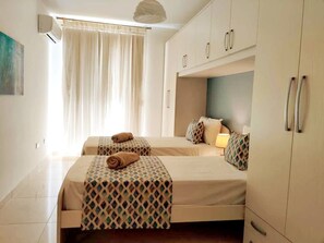 A hotel room with two beds, a window with curtains, a ceiling fan, and a minibar. - GrandBlue Vista Premier Seafront Apartment (Żebbuġ)
