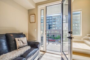 Ein modernes Wohnzimmer mit einem schwarzen Ledersofa, einer weißen, flauschigen Decke und einer Glastür, die auf einen Balkon mit Blick auf die Stadt führt.