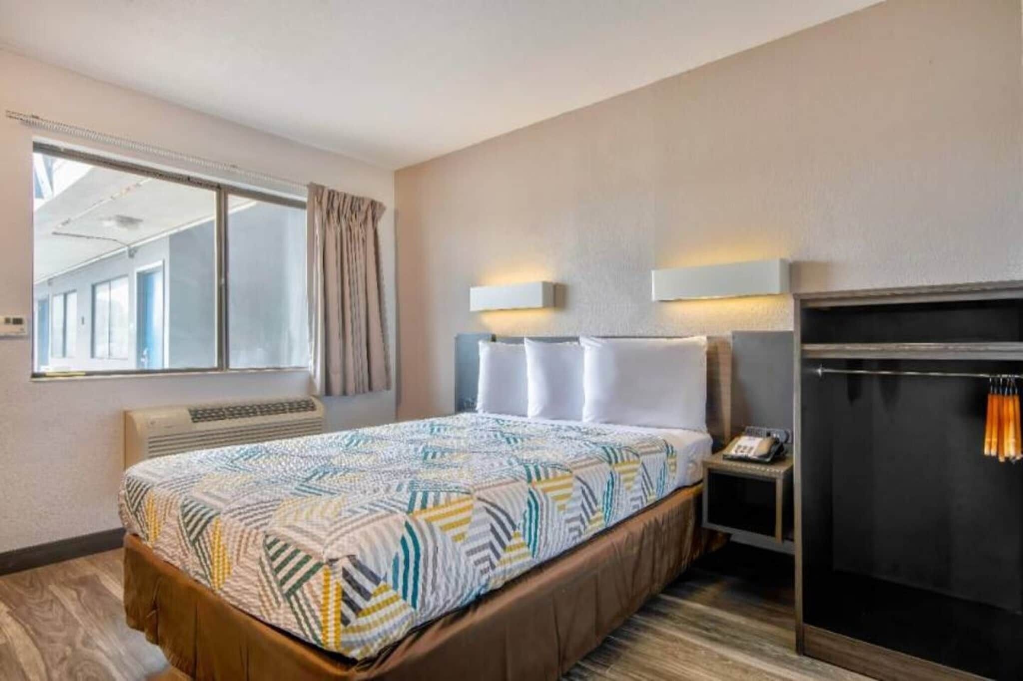 A hotel room with a bed, a bedside table with a phone, a heater, a window with curtains, and a wall-mounted light fixture.