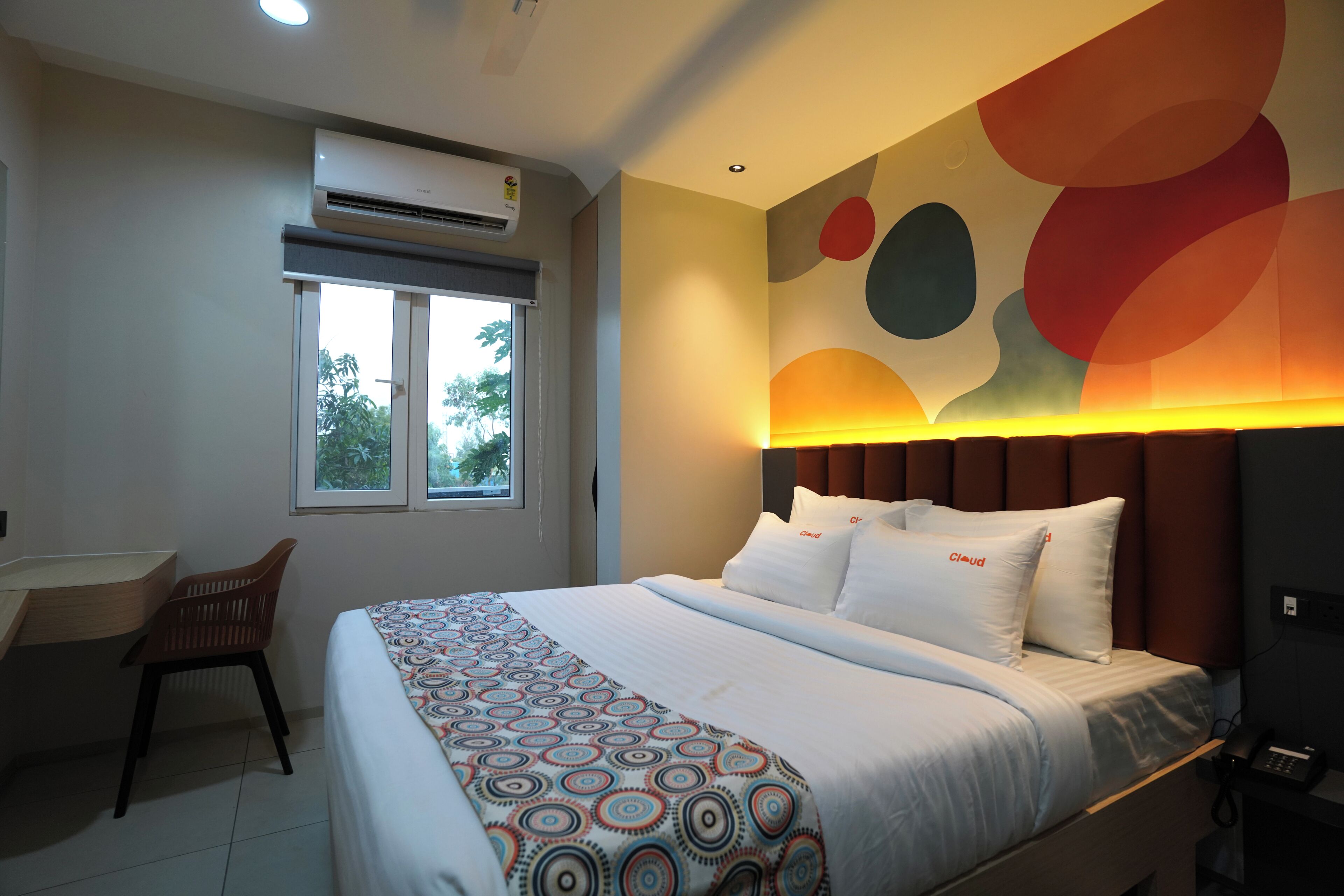 A hotel room with a bed, a desk, a chair, an air conditioner, and a window.