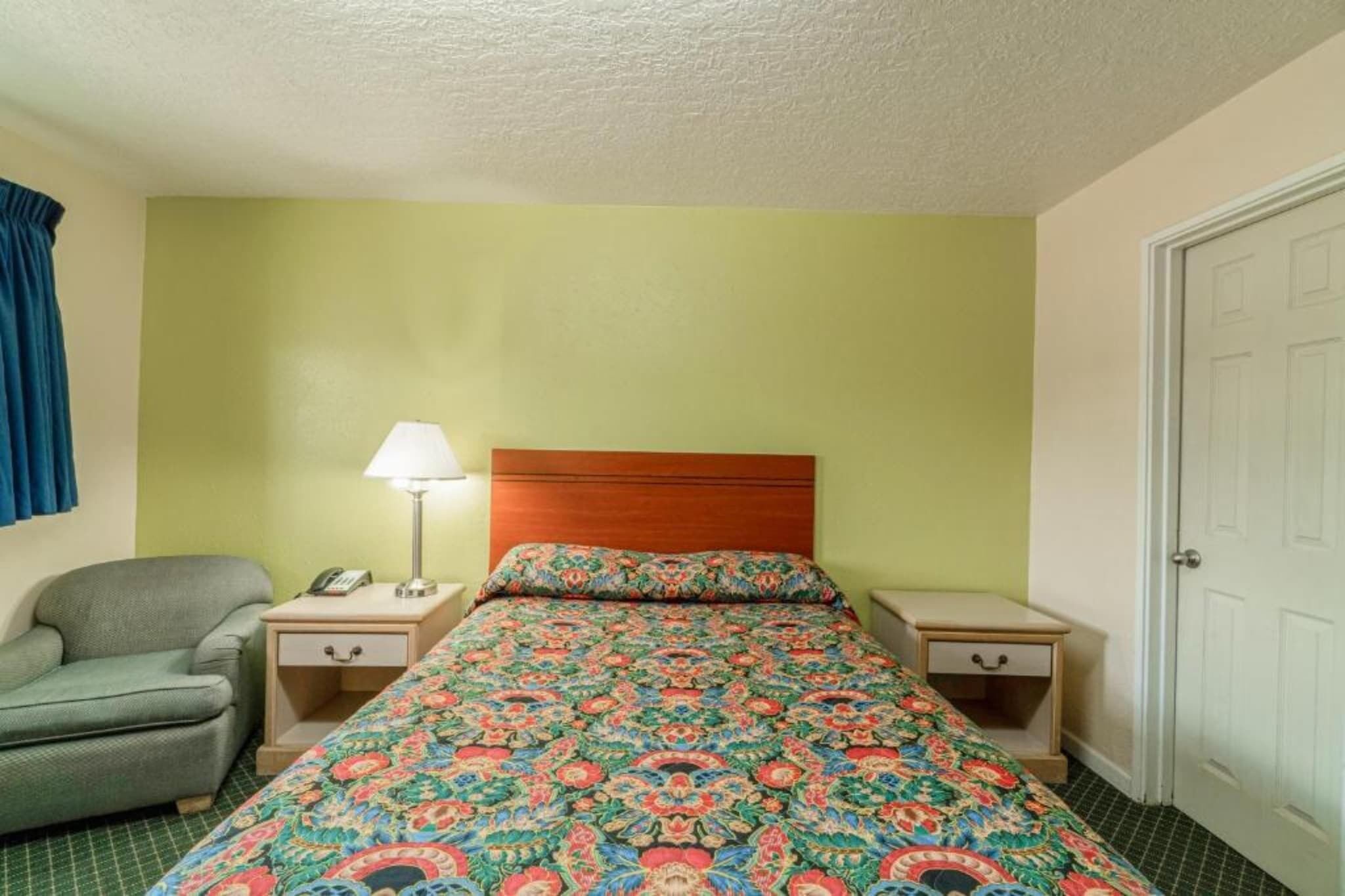 Una cama individual con una colcha estampada, una lámpara de noche, una silla, una mesita de noche con un teléfono y una puerta.