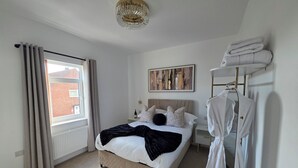 A bedroom with a bed, a hanging robe rack, a painting, and a window with curtains. - Newly Refurbished
Suburban Luxe By Luxe Sleep (Stockport)