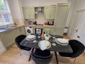 Mesa de comedor para dos personas con cubierta de mármol negro, rodeada de sillas negras, en una cocina con armarios verde claro y una ventana.