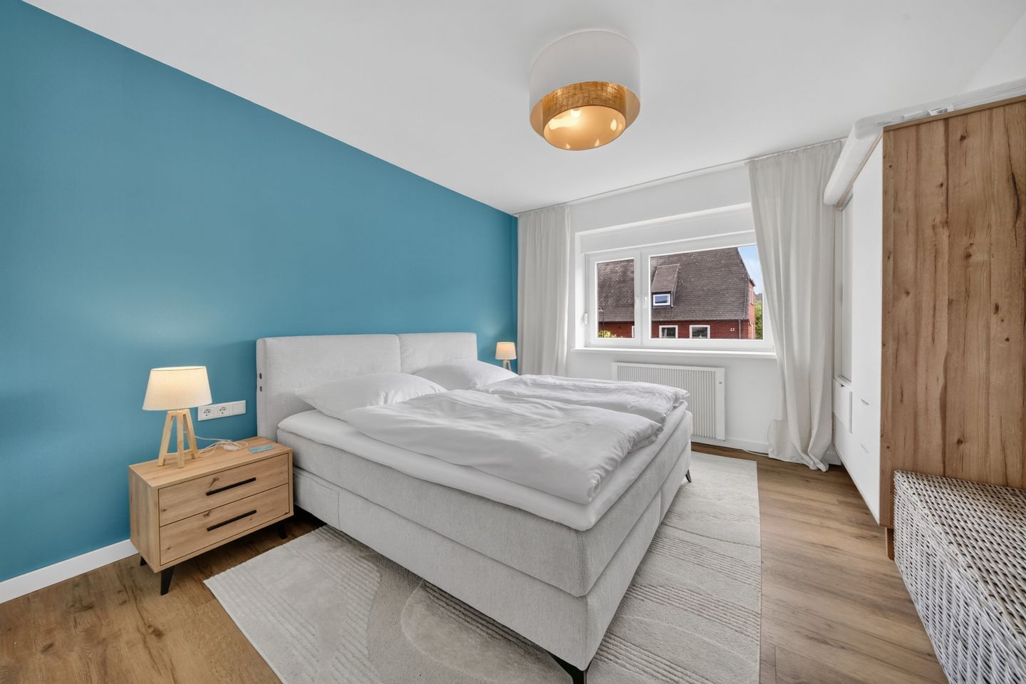 Een slaapkamer met een bed, nachtkastje, lamp, raam en uitzicht op een gebouw.
