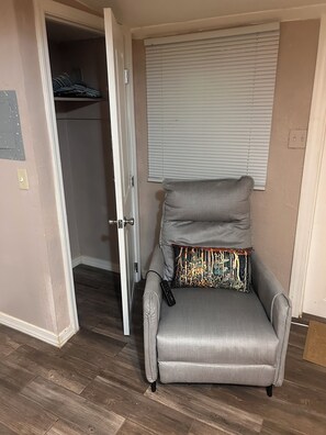 A grey armchair with a patterned cushion, a remote control on it, and a closet with clothes hanging in the background.