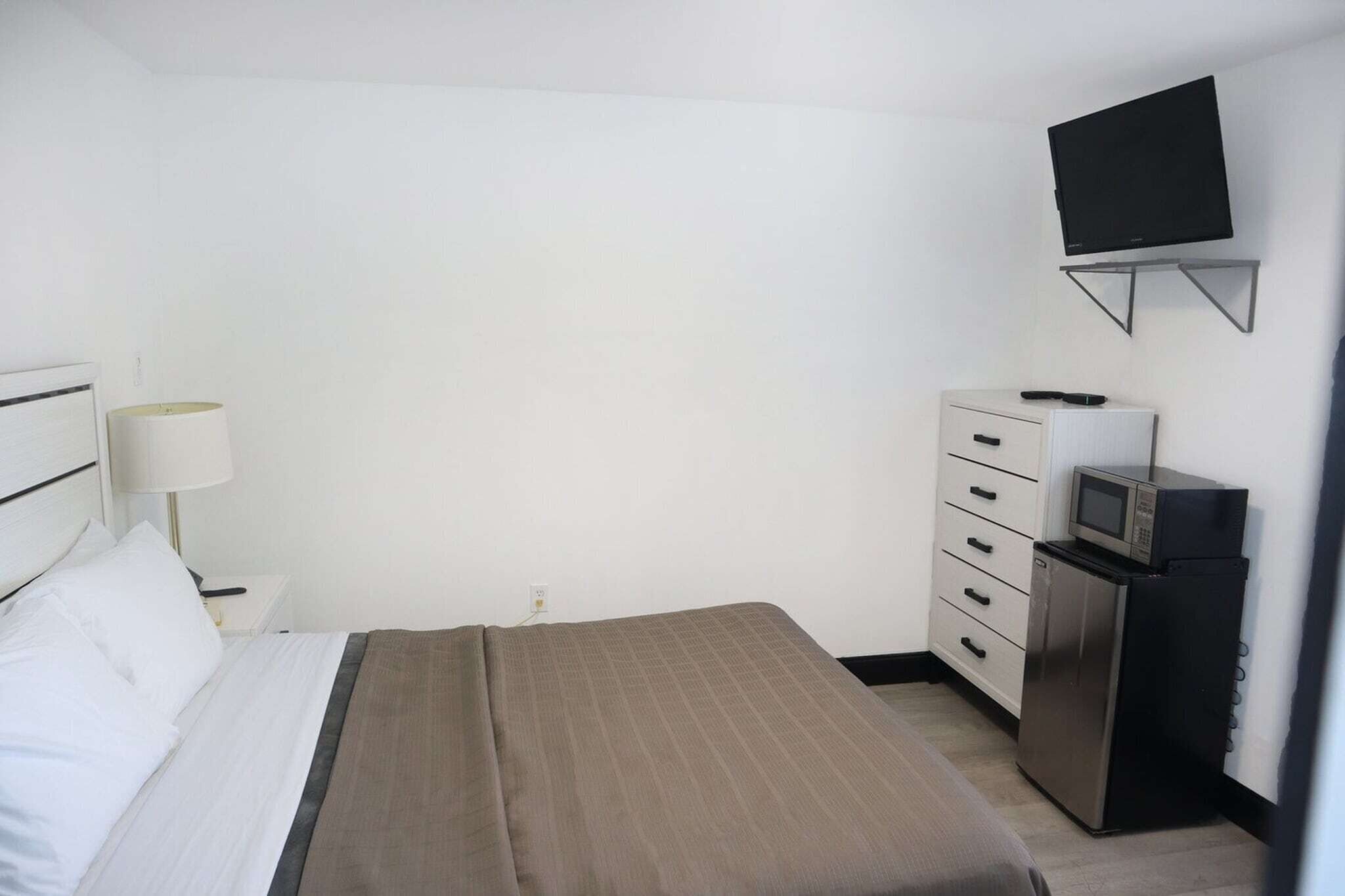 A hotel room with a bed, a TV mounted on the wall, a desk with a lamp, and a refrigerator.