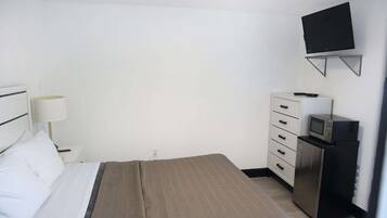 A hotel room with a bed, a TV mounted on the wall, a desk with a lamp, and a refrigerator.