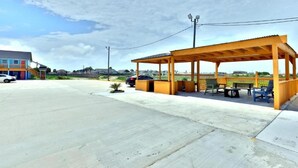 Een overdekt terras met een houten constructie, een verharde parkeerplaats en een gebouw op de achtergrond.
