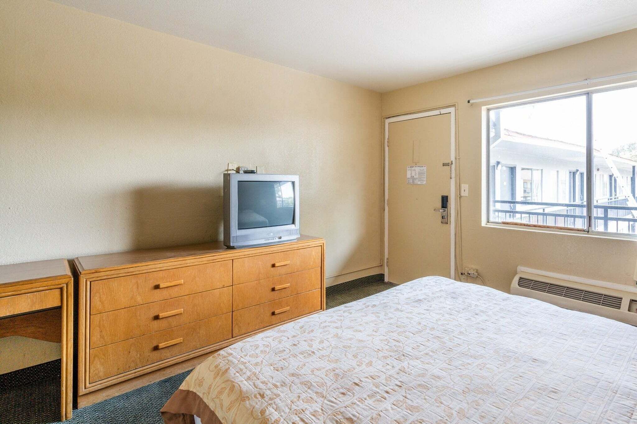 A hotel room with a bed, a wooden dresser, a television on the dresser, a window, and a door.