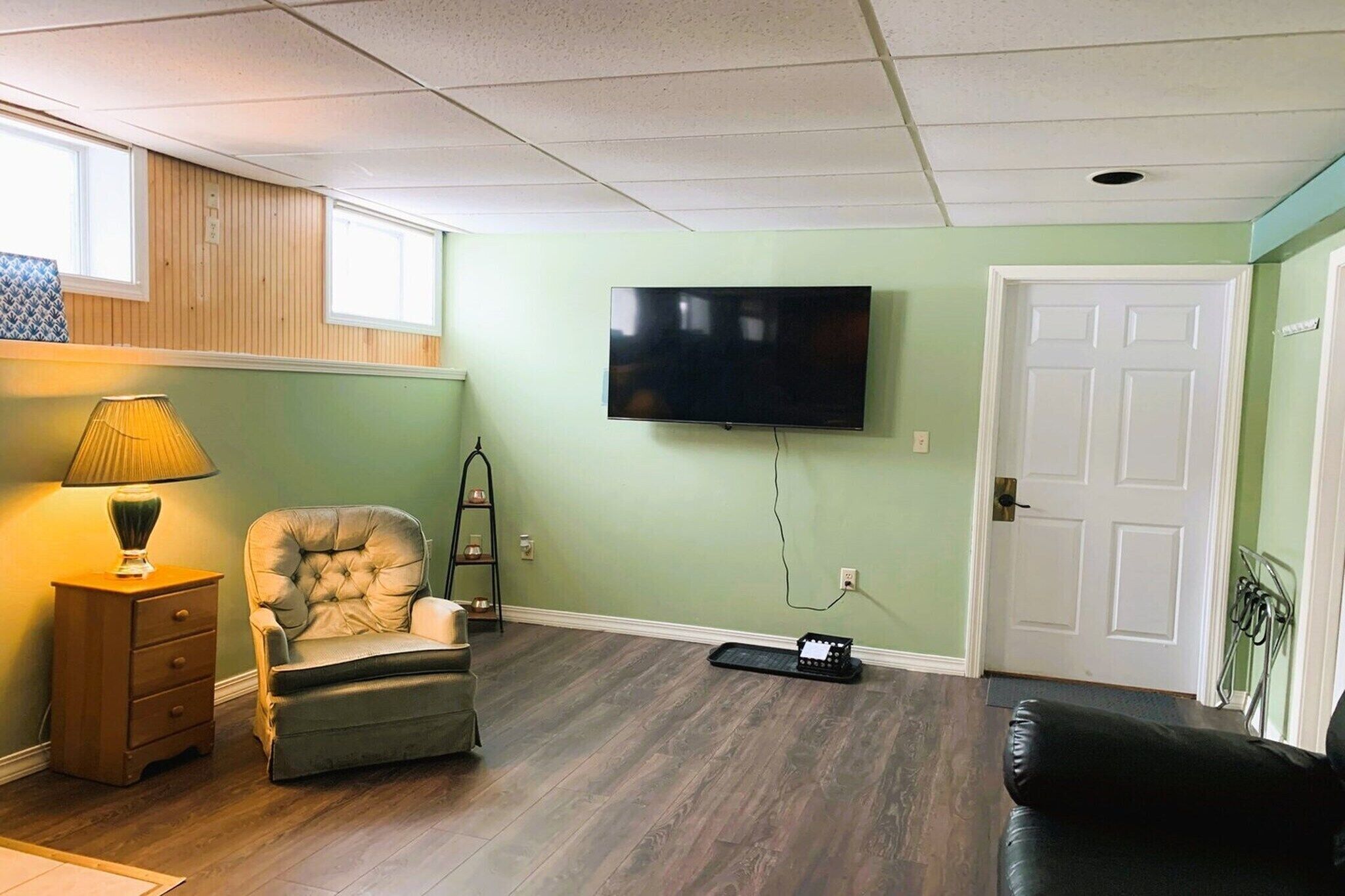A living room with a flat-screen TV mounted on the wall, a leather armchair, a wooden nightstand with a lamp, and a door with a metal handle.