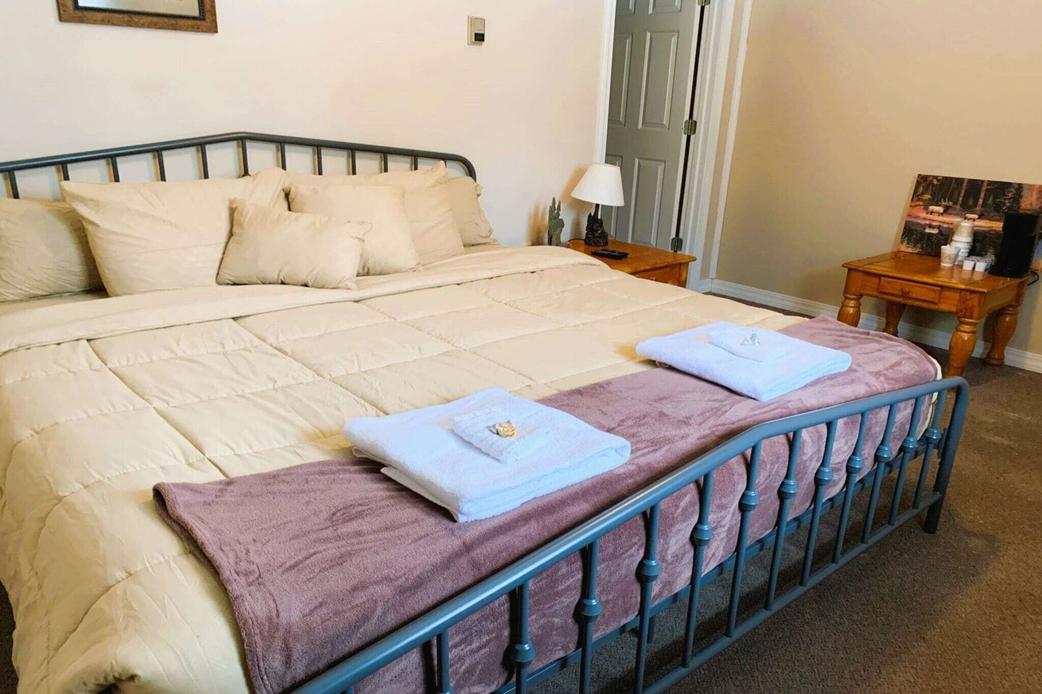A bed with a quilted cover, two folded towels, a bedside table with a lamp, and a small wooden table with items.