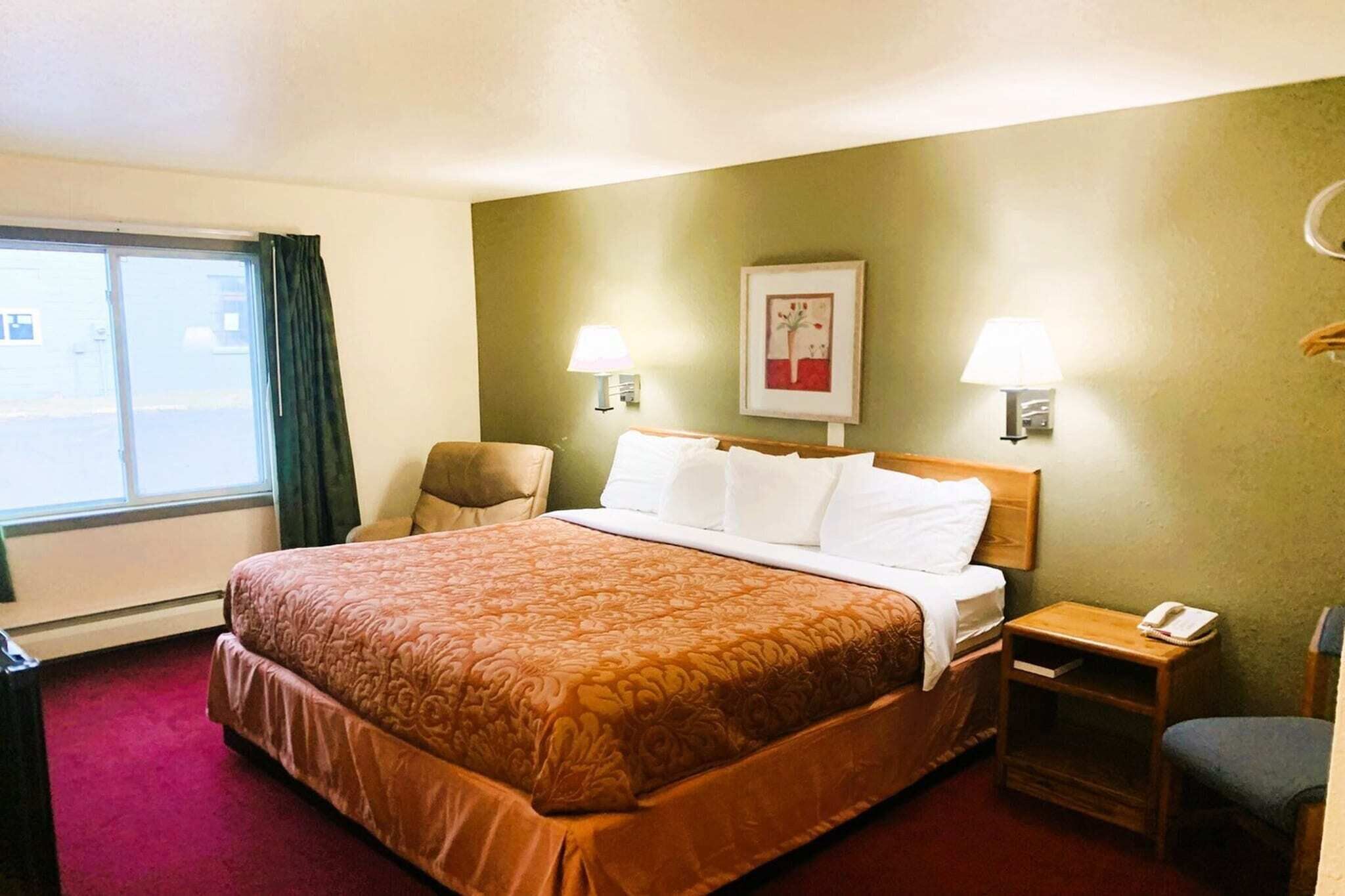 A hotel room with a large bed, two bedside lamps, a chair, a small table with a phone, and a window with curtains.