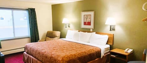 A hotel room with a large bed, two bedside lamps, a chair, a small table with a phone, and a window with curtains.