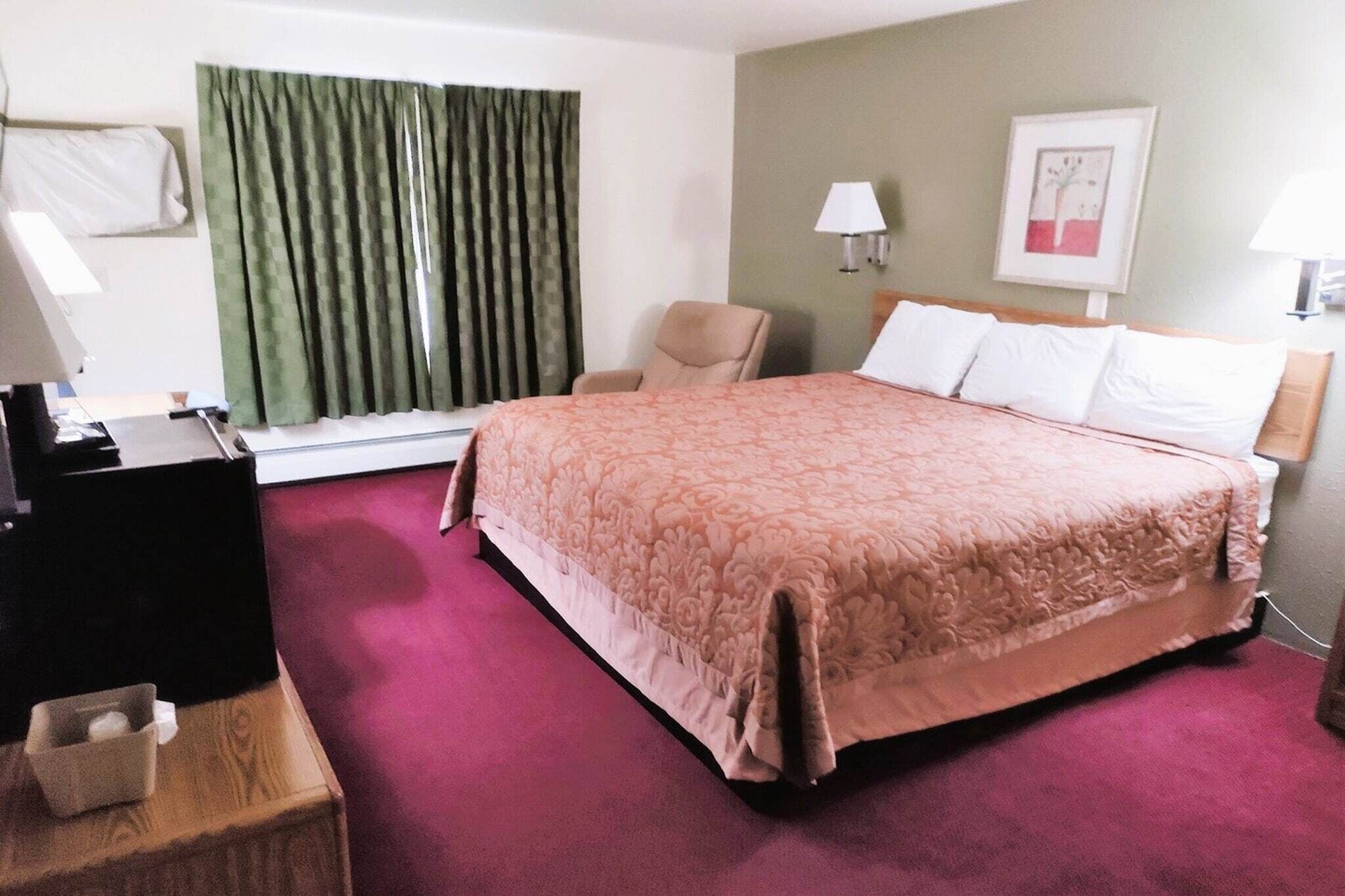 Quarto de hotel com uma cama grande, uma cadeira, uma escrivaninha e uma janela com cortinas.