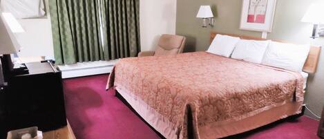 Quarto de hotel com uma cama grande, uma cadeira, uma escrivaninha e uma janela com cortinas.