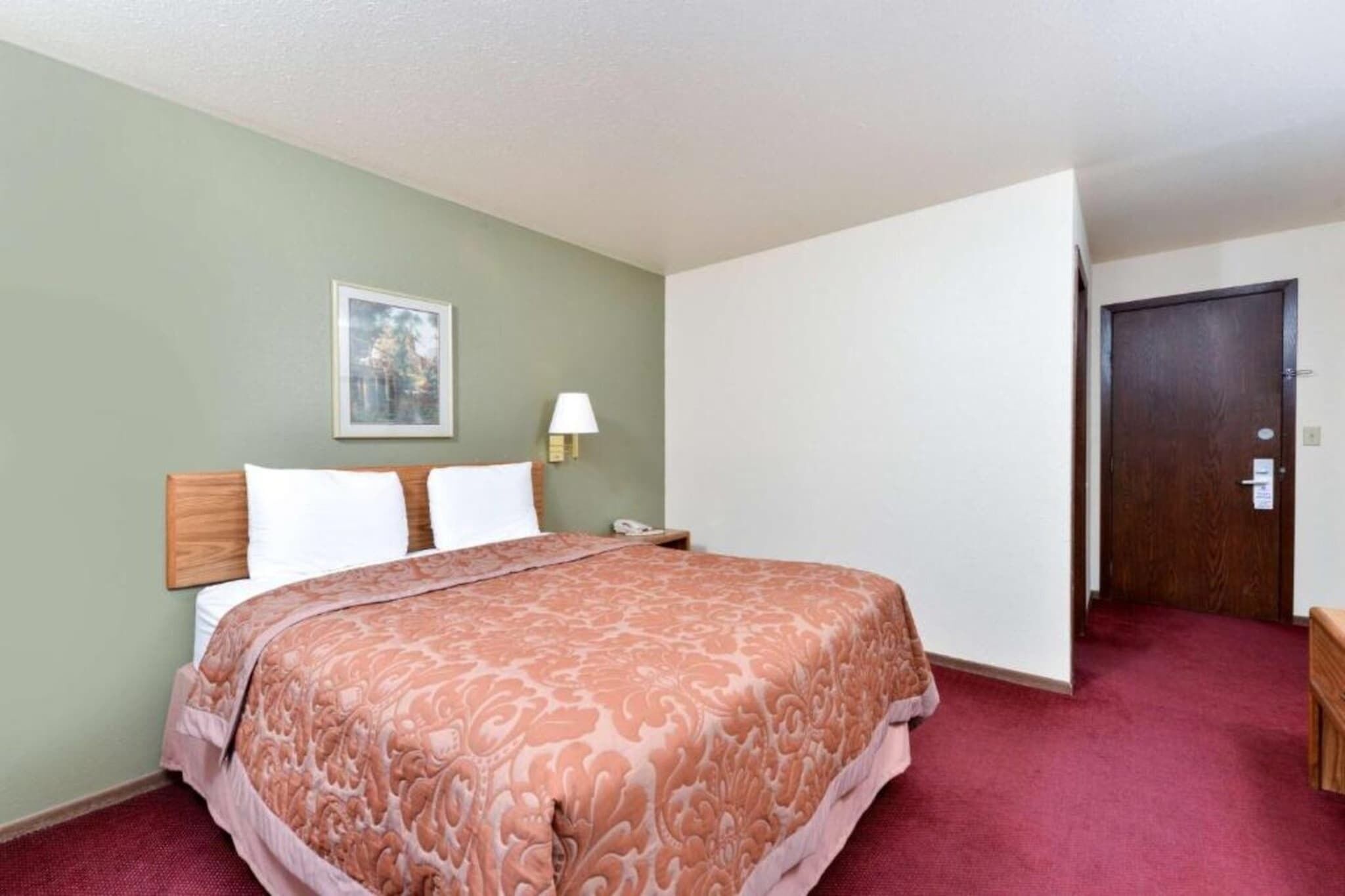 A hotel room with a bed, a nightstand, a lamp, a door, and a framed picture on the wall.