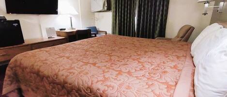 A hotel room with a large bed, a desk with a lamp, a television, and a chair.