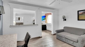 A spacious living area with a ceiling fan, a gray sofa, a dining table, and a kitchen in the background.