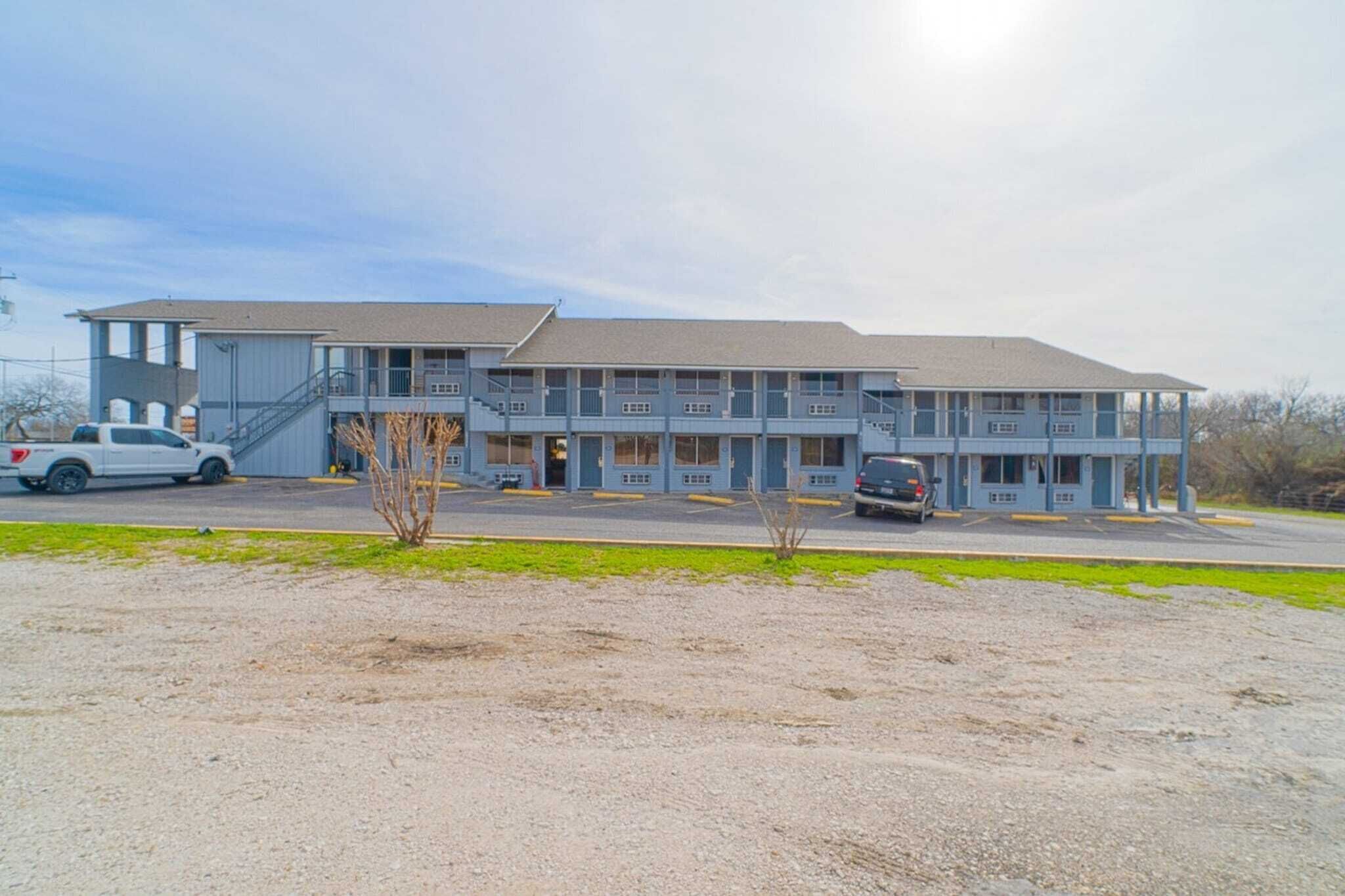 Un motel à plusieurs étages, doté d’un toit plat, d’un espace de stationnement couvert et d’un parking en gravier à l’avant.