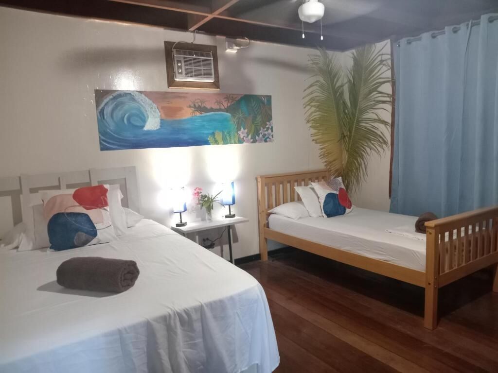 A room with two beds, a desk with a lamp, a large plant, and a painting on the wall.