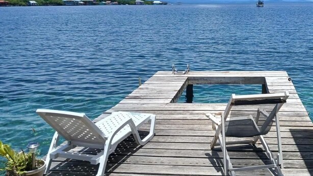 Un muelle de madera con dos sillones blancos y una pequeña mesa, con vista a un mar tranquilo y un velero a lo lejos.