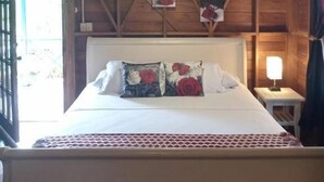 Techo de madera, una cama con almohadillas florales, una lámpara de noche y una ventana con cortinas.