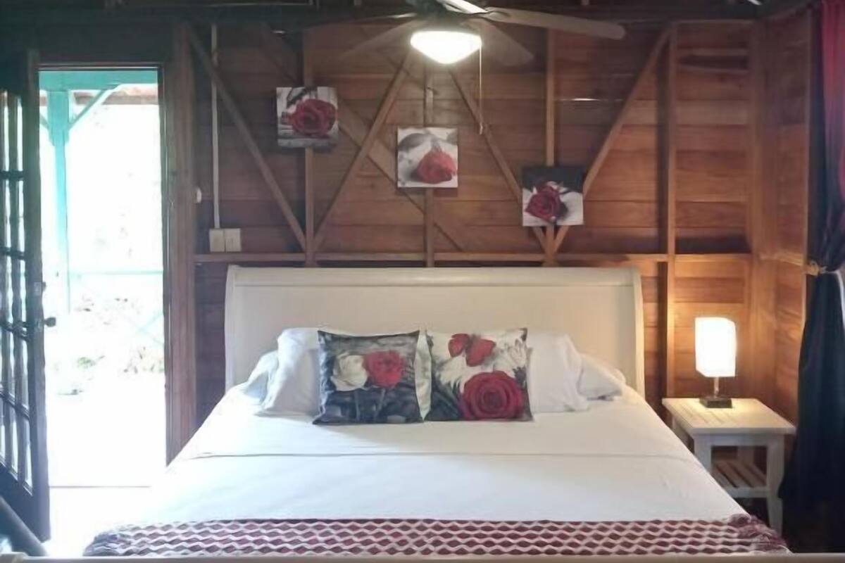 A wooden ceiling, a bed with floral pillows, a bedside lamp, and a window with curtains.