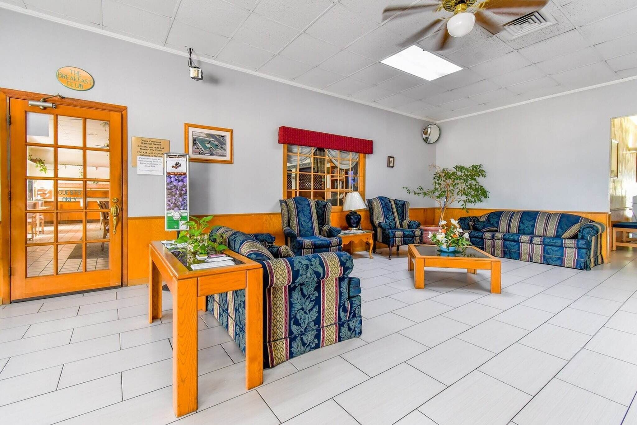 A hotel lobby with a reception desk, seating area with sofas and chairs, a coffee table, a clock, and a sign indicating a breakfast club.