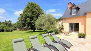 Un patio con cuatro sillones de descanso, una casa con techo inclinado y un árbol grande al fondo.